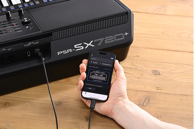Yamaha 數位工作站 PSR-SX720+ 與手持顯示 Yamaha 官方應用程式「Expansion Explorer」畫面之智慧型手機的照片。